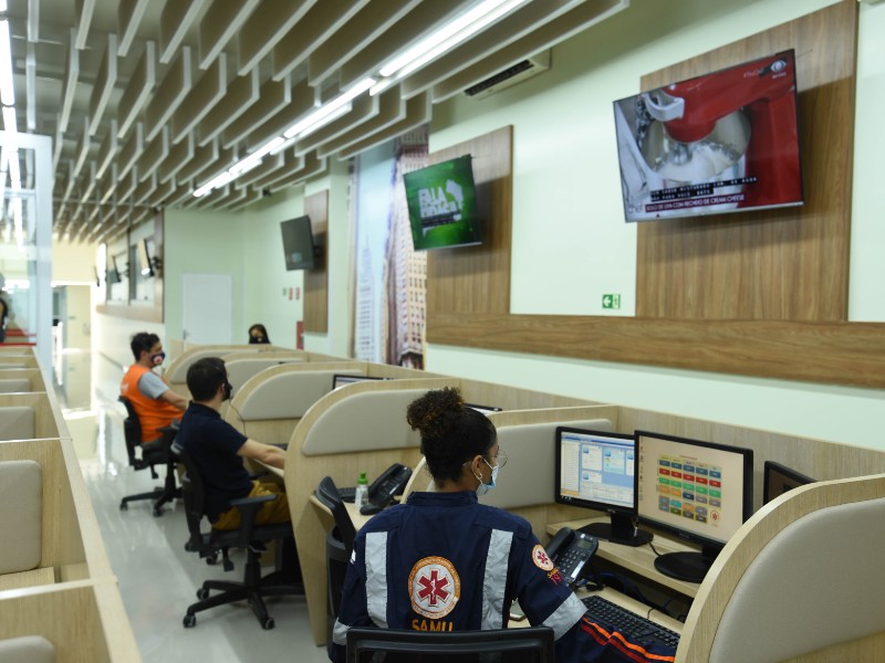Quatro pessoas trabalham numa bancada de madeira com divisórias e equipadas com computador e telefone. Na parede, há três televisores 